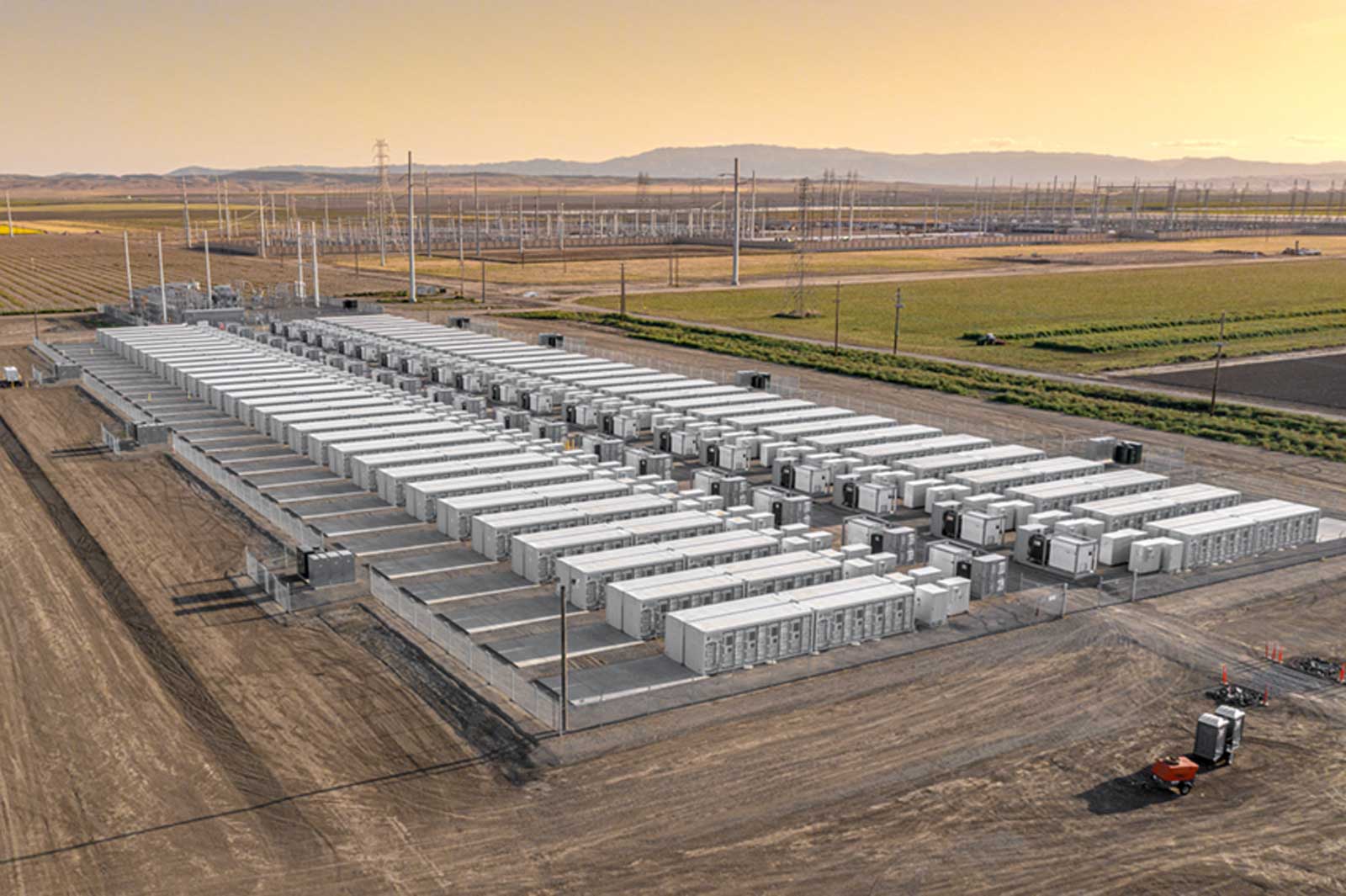 A large utility-scale battery energy storage facility sits on a cleared, gravel-covered site in a rural landscape. Dozens of long, white container-like battery units are arranged in neat parallel rows, each connected by electrical equipment and fenced for security. Power lines and a large substation stretch across the background, with farmland and low hills visible under a golden sky.
